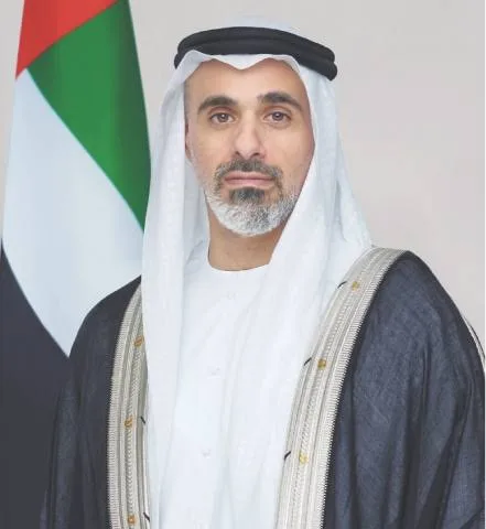 Portrait of Sheikh Khaled bin Mohamed bin Zayed Al Nahyan in a white ghutra and dark bisht, standing in front of the flag of the United Arab Emirates