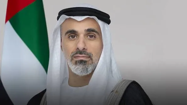 Portrait of Sheikh Khaled bin Mohamed bin Zayed Al Nahyan in a white ghutra and dark bisht beside the flag of the United Arab Emirates